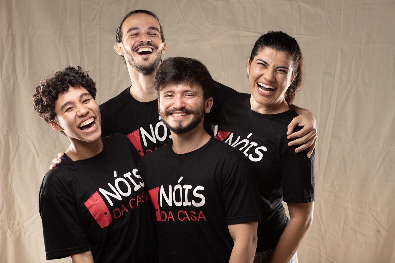 Quatro integrantes do Nóis da Casa reunidos, sorrindo e usando camisetas pretas com a logo do grupo